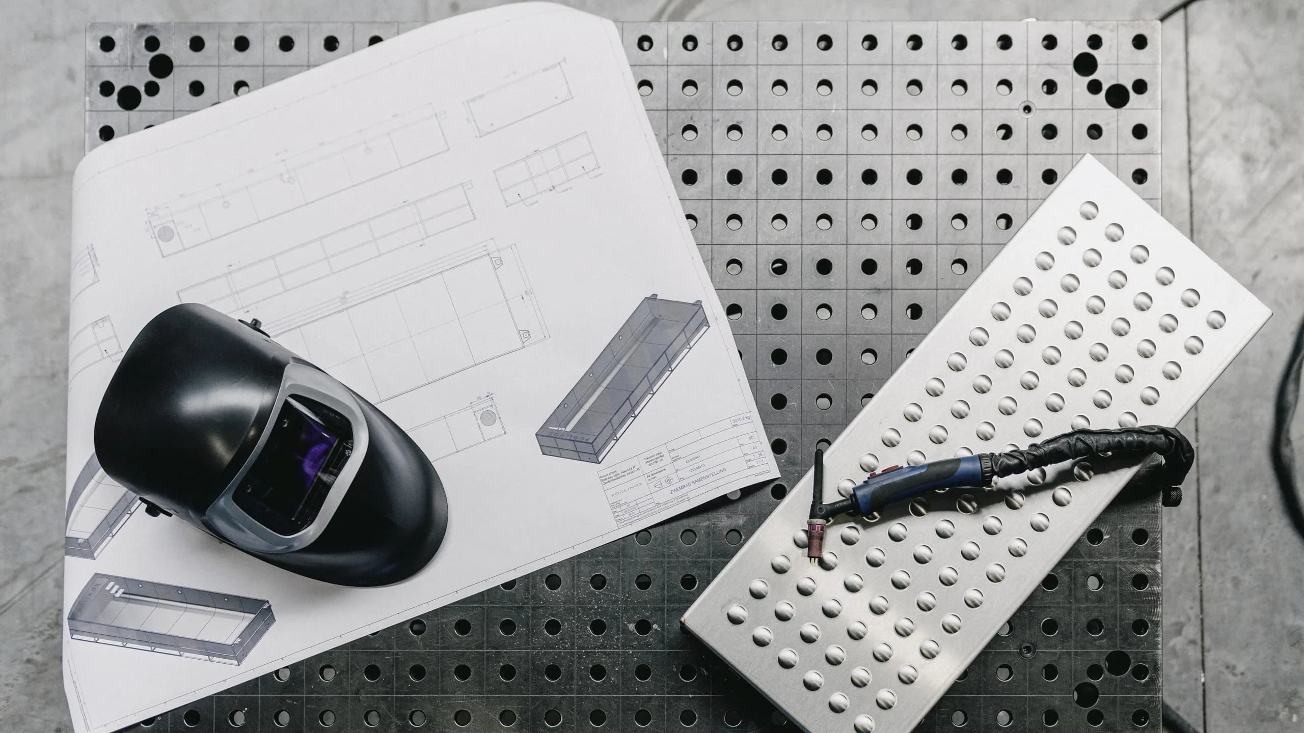Welding helmet, technical drawings and a stainless steel pool component on a workbench